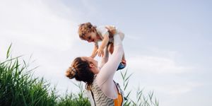 woman holding laughing baby up in the air stood in grass